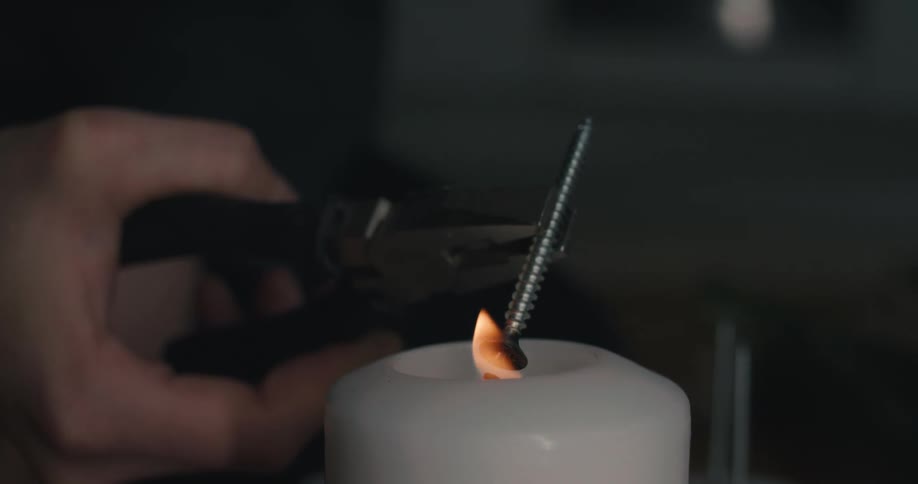 An adult hand uses a pair of pliers to hold a metal screw against a candle flame