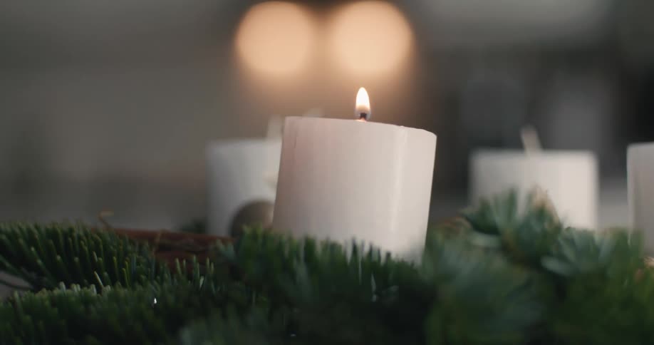 Nestled among evergreen branches a white pillar candle burns brightly its flame dancing softly before a backdrop of warm outoffocus golden lights