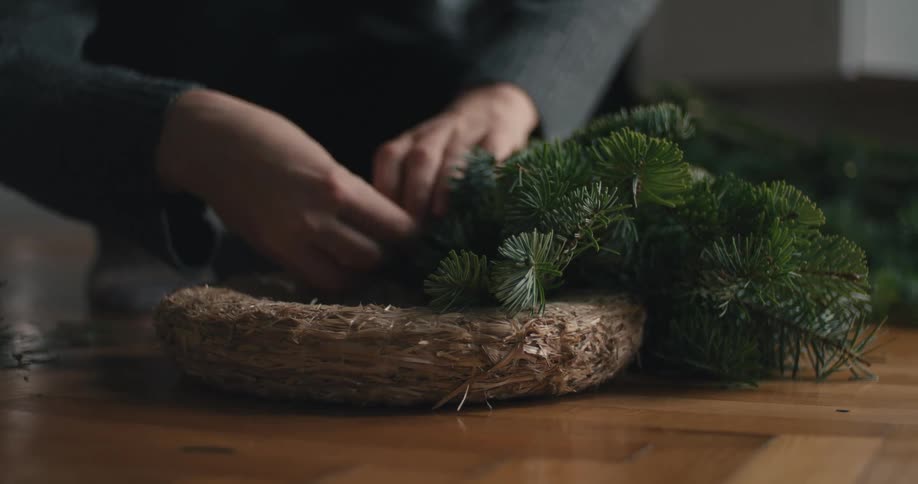 A persons hands wearing a dark sweater work to attach fresh evergreen branches to a circular straw base crafting a holiday wreath on a wooden surface in a lowlight setting