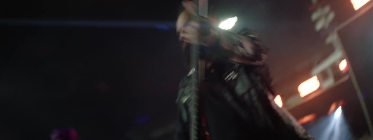 A close quickmoving shot shows a bearded musician wearing a dark leather jacket aggressively strumming a black guitar on a dark dimly lit stage during a concert performance and a female guitarist on the back