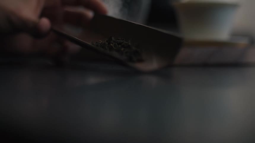 A persons hand carefully uses a dark wooden scoop to move a small pile of dried green tea leaves on a reflective surface with softfocus tea ware visible in the background