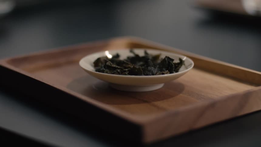 A small shallow white ceramic dish containing dark coiled tea leaves rests on a rectangular wooden tray on a dark surface