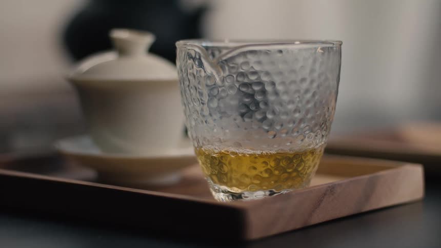 This video displays a closeup of a small glass pitcher with a textured bubbled surface containing a small amount of goldenyellow liquid likely tea the pitcher is sitting on a wooden tray alongside a white lidded teacup and saucer in the softly lit