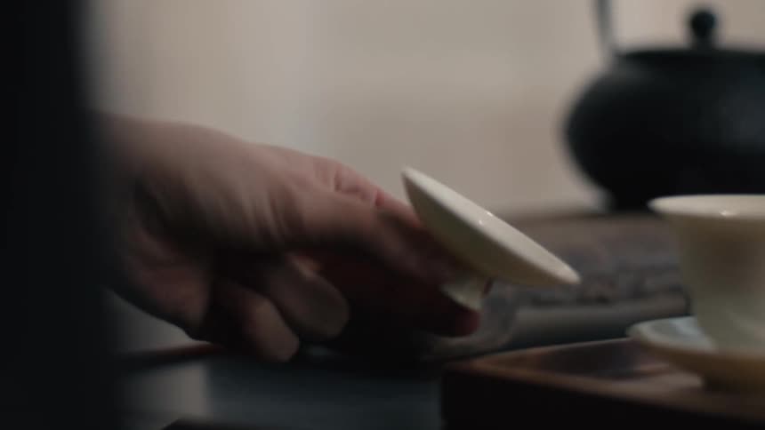 The video focuses on a clear textured glass pitcher containing ambercolored tea sitting on a wooden serving tray alongside a traditional white teacup and saucera hand gently places a small round white lid next to the teacup with a dark teapot visi