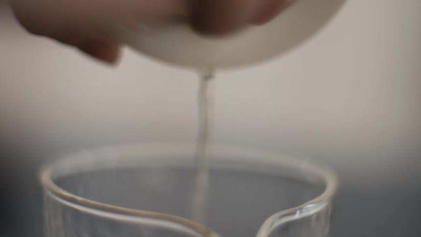 A closeup shot of a hand holding a white ceramic vessel pouring a stream of clear liquid into a glass measuring beaker ending the pour