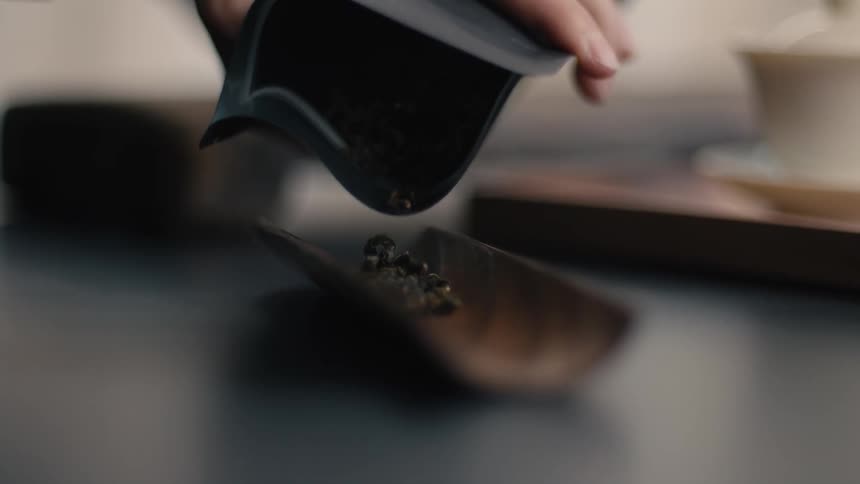 A person pours dark green dried tea leaves from a black pouch onto a shallow wooden scoop resting on a dark surface in a close view