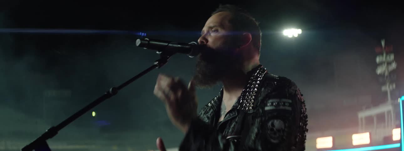 A man with a beard and tattoos wearing a studded leather jacket sings loudly into a microphone on a stage in a dark smoky stadium concert