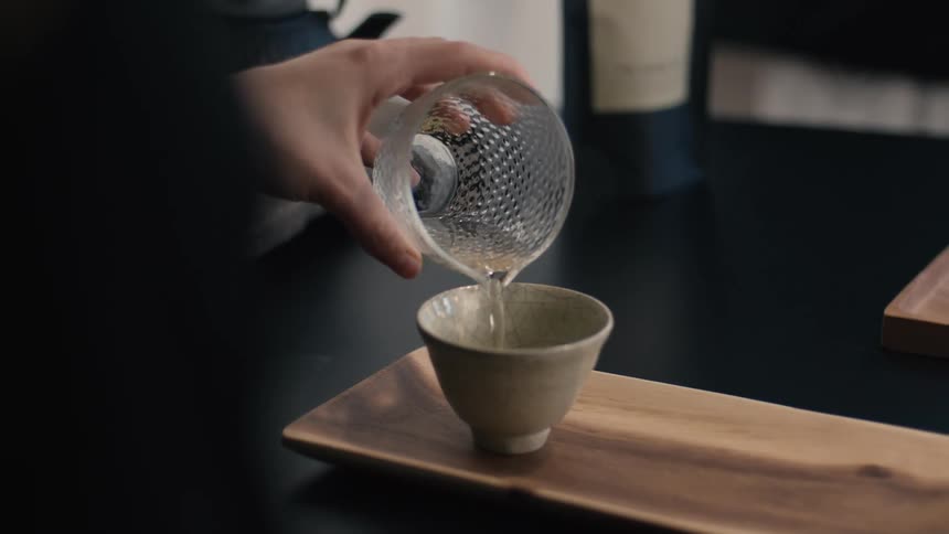 Male adult hands pouring a beverage from a glass cup into a ceramic bowl over a wooden tray on a table indoors