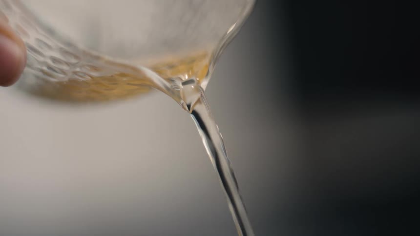 A very closeup shot captures a person precisely pouring a steady clear stream of goldenamber colored liquid presumably tea from a textured glass pitcher into a small cracked ceramic cup