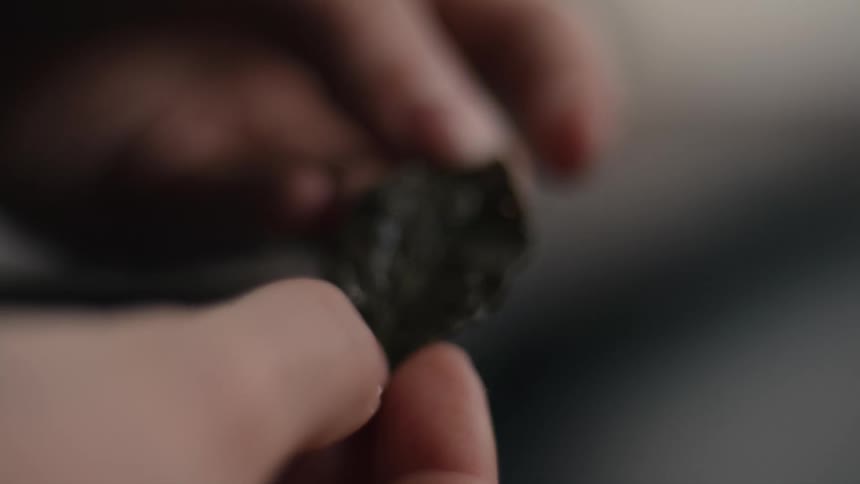 A person is shown holding a small rough dark gray rock or mineral fragment between their fingers closely examining its surfacethe camera focuses tightly on the stone highlighting its irregular somewhat glossy texture as it is slightly turned in th
