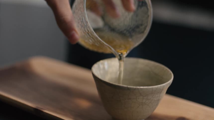 A persons hand pours a stream of lightcolored liquid from a clear textured glass pitcher into a small cracked ceramic cup resting on a simple wooden tray