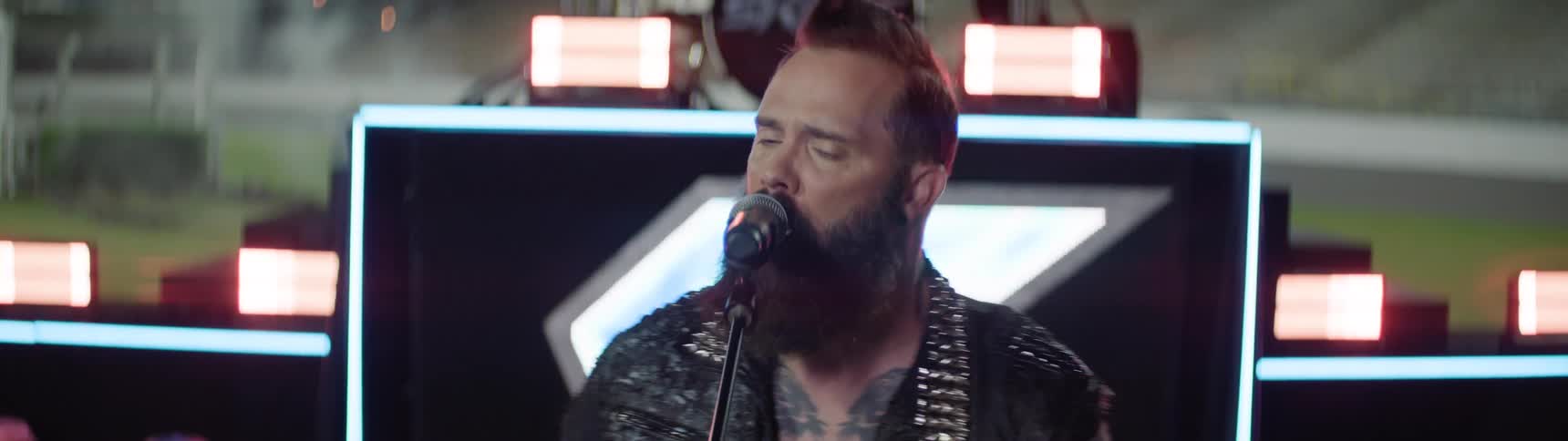 A bearded rock singer in a studded leather jacket performs passionately into a microphone on a lit stage