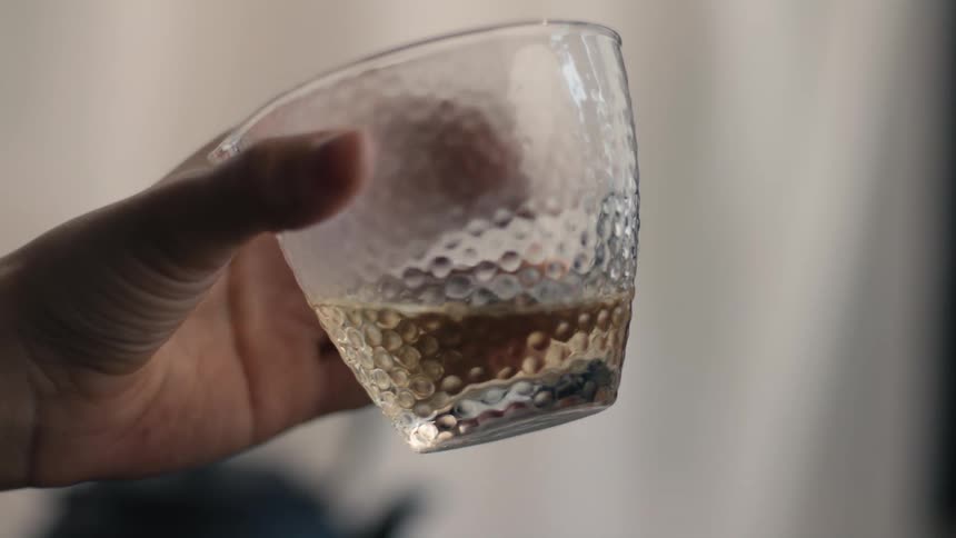 A womans hand holds a textured clear glass cup filled with light amber liquid gently tilting the cup against a soft indoor background
