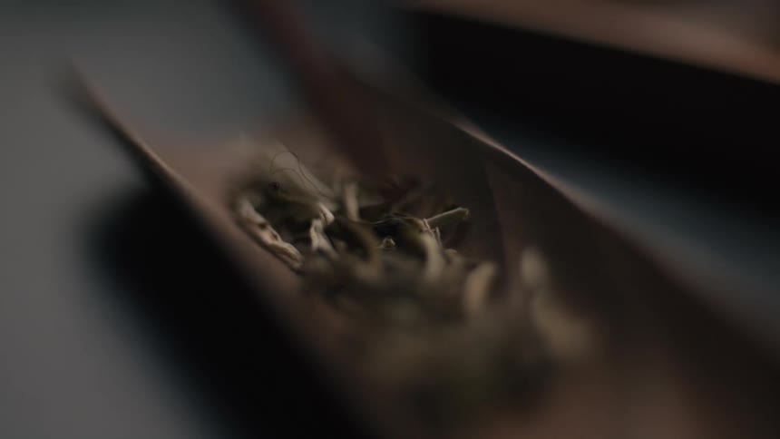 A closeup shot focuses on dry light green and white loose leaf tea being gently pushed or poured from a curved wooden scoop onto a surface in a dimly lit dark setting