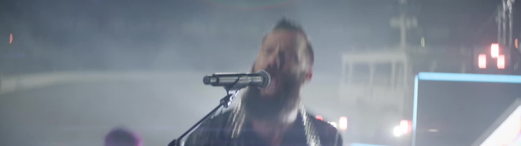 This is a video clip of a band performing live on stagea bearded male musician with a dark outfit is playing a guitar or bass moving energetically under bright stage lights and heavy fogsmokeanother band member possibly with purple hair is brie
