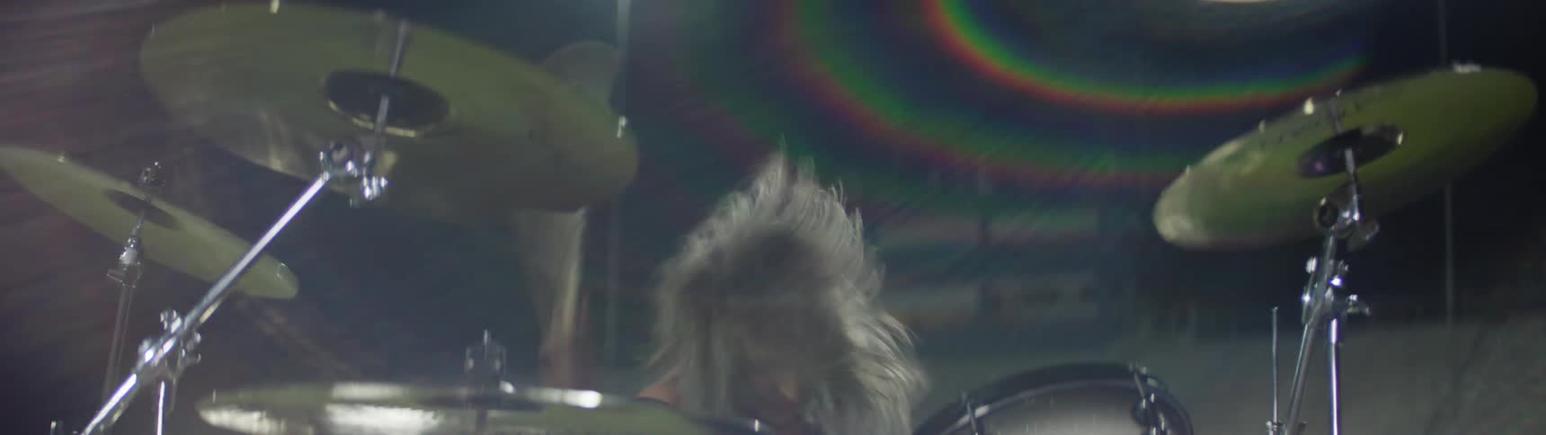 A closeup captures a female drummer with flowing gray hair vigorously striking the drum kit and cymbals during an energetic nighttime performance illuminated by bright stage lights