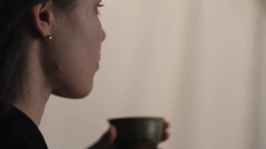 A woman in profile lifts a small dark ceramic cup to her lips taking a drink in a dimly lit indoor space