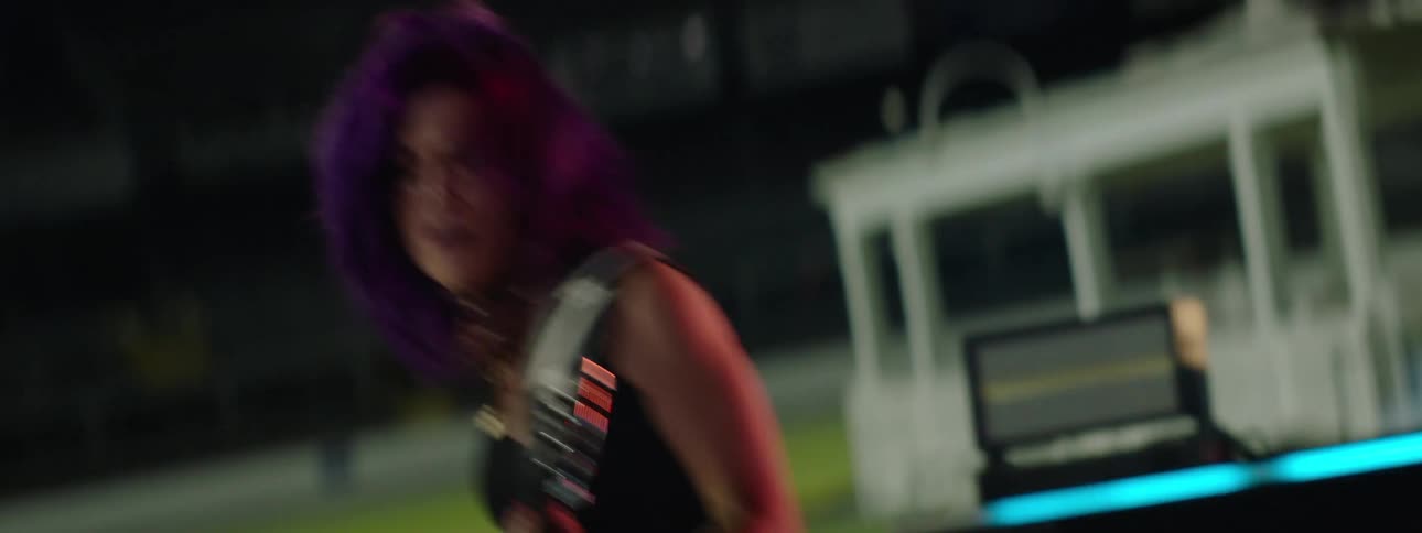 A bearded man sings on a microphone next to a woman with vibrant purple hair wearing a dark sleeveless top vigorously plays an electric guitar on a brightly lit stage