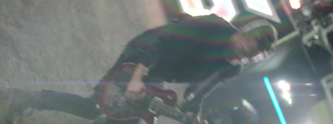 An intense performance featuring a guitarist and other band members rocking out on a stage framed by large brightly lit geometric props with a drummer positioned on a riser in the background