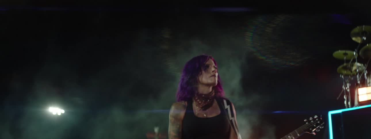 A woman with purple hair and tattoos plays an electric guitar on a dimly lit outdoor stage with bright neon blue lights