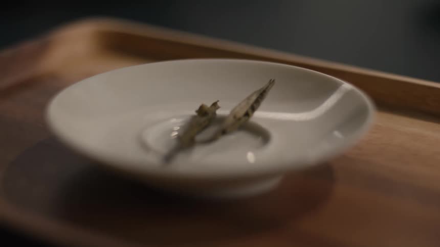 A small white ceramic dish holds a few delicate pale green dried tea leaves on a smooth wooden serving tray in a dim interior