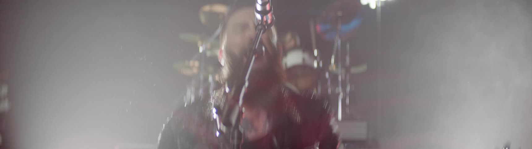 Bearded man playing an electric guitar and singing into a microphone on a stage at a concert with other band members around him indoors