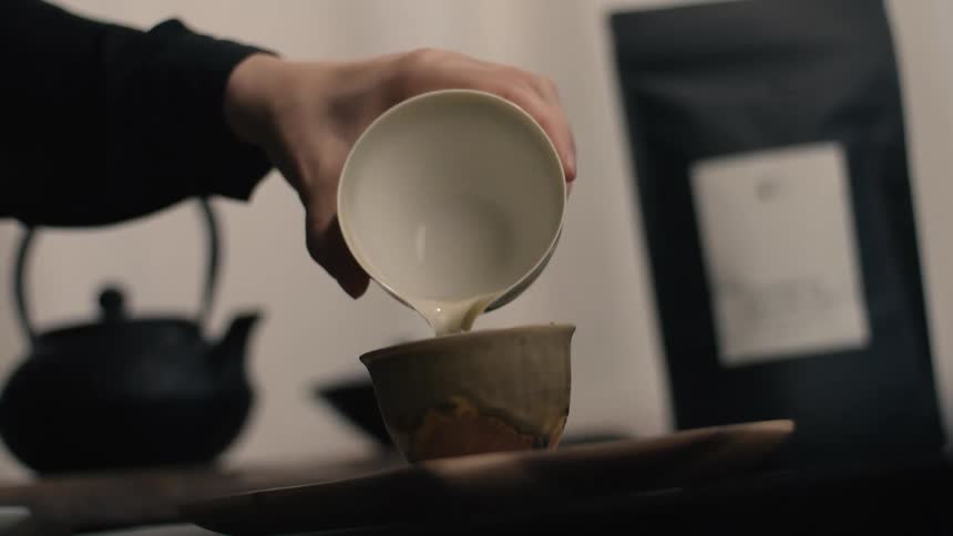 A hand pours light amber tea from a white ceramic vessel into a small rustic cup on a wooden board next to a black teapot