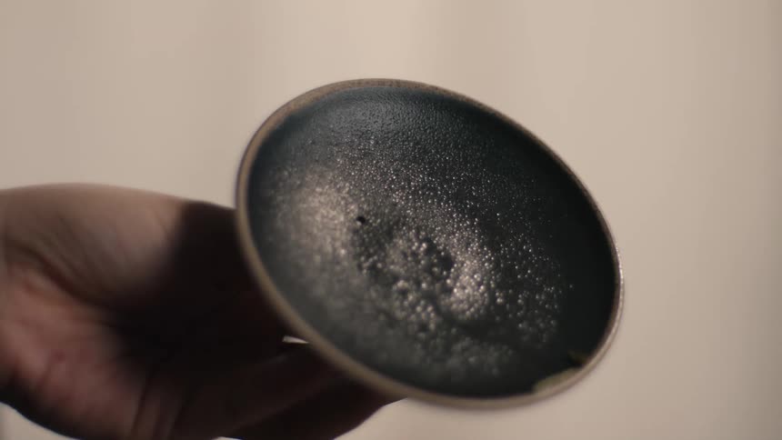 A hand carefully transfers the final tea leaves from an inverted dark small bowl into a similar receiving bowl completing the precise action of portioning the tea on a wooden tray with a glass vessel nearby