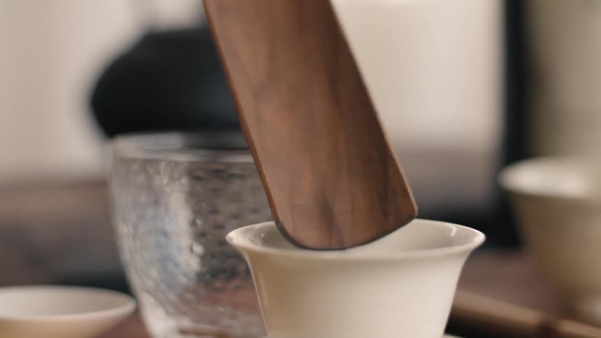 A closeup shot captures a hand using a small wooden scoop to pour a portion of dark dry tea leaves into a delicate white ceramic cup on a table