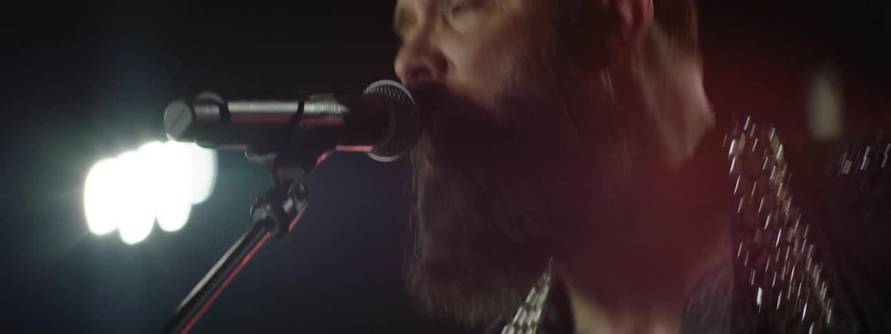 Bearded singer in patterned shirt with hand tattoos sings intensely into microphone under dramatic lighting