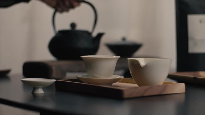 Male hands pouring hot water from a black kettle into a white ceramic cup on a wooden tray over a table indoors