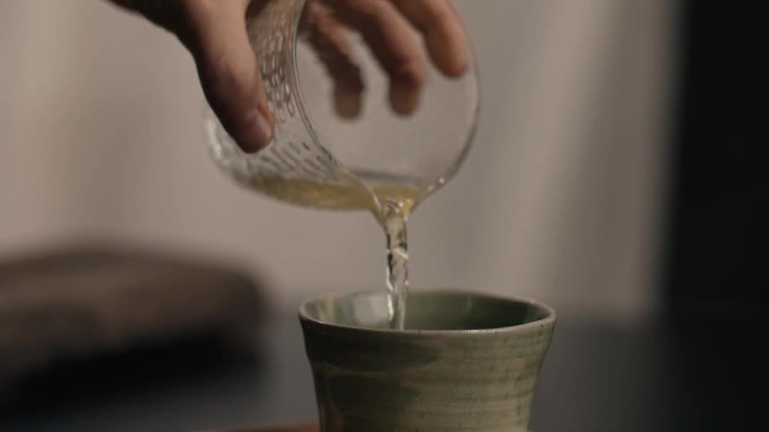 Person pouring a yellow beverage from a glass cup into a ceramic cup indoors