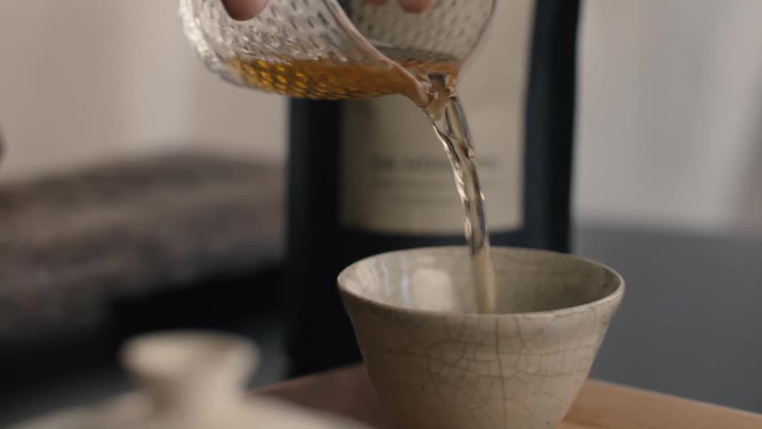 A hand pours amber tea from a textured glass pitcher into a small ceramic cup