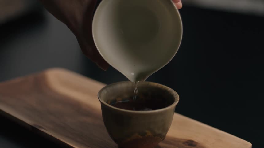 A hand carefully pours light colored tea from a white ceramic vessel into a small dark brown cup resting on a wooden tray