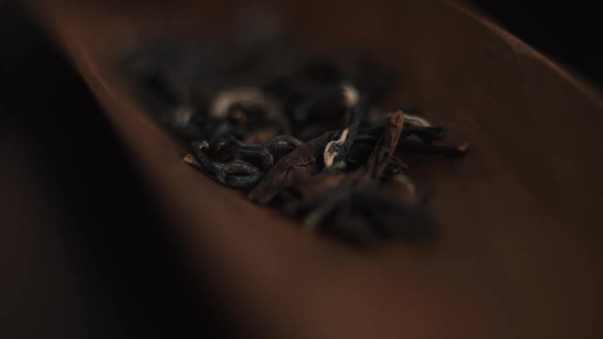 A very close view of dark dried tea leaves resting on a shallow wooden scoop in a dark atmospheric setting