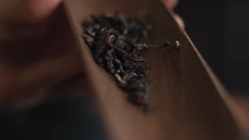 A closeup shot of a persons hands using a wooden scoop to gently pour dry dark tea leaves into a small white ceramic cup initiating a traditional tea preparation ritual