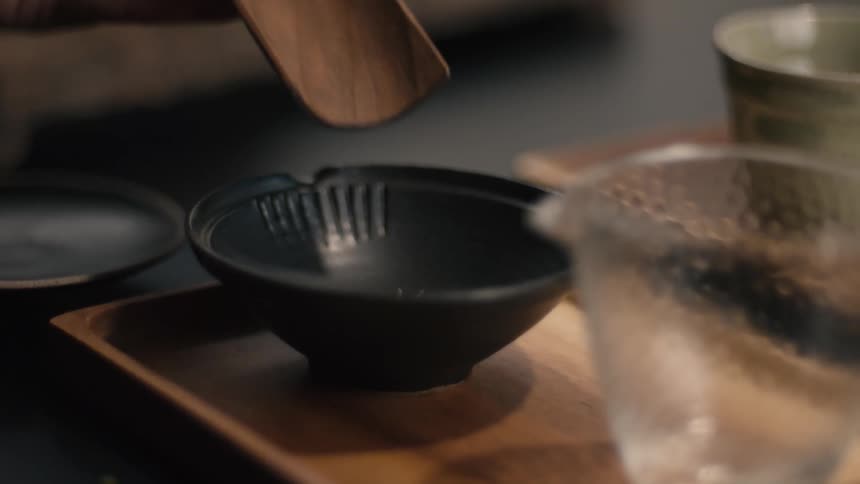 A hand gently pours loose tea leaves from a wooden scoop into a small dark bowl