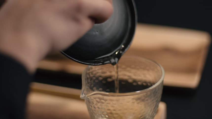 Liquid being poured from dark ceramic vessel into textured glass pitcher on wooden surface