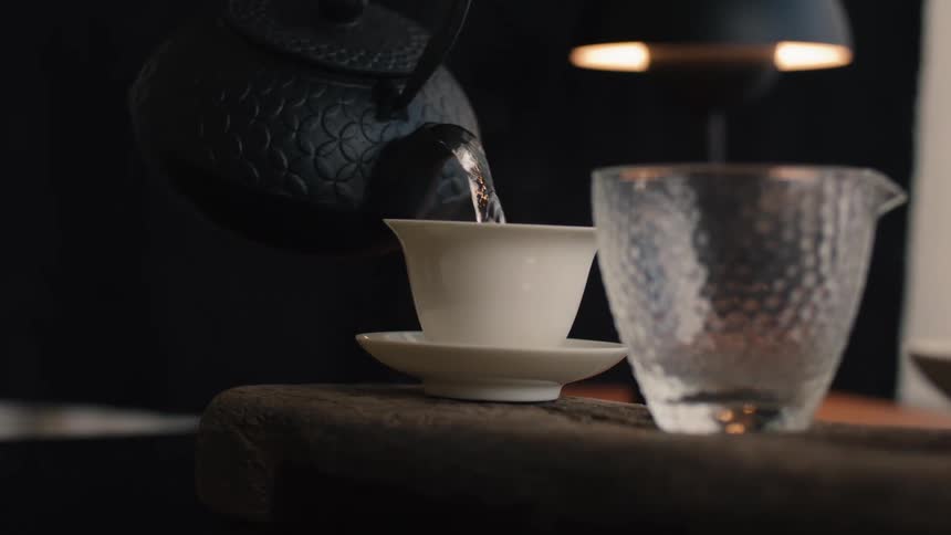 A closeup shot captures a hand pouring steaming hot water from a black textured cast iron teapot into a small white ceramic teacup sitting on a saucer during a tea preparation