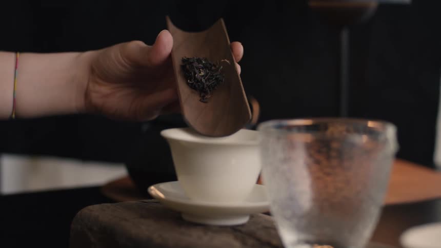 Human hands putting tea leaves into a ceramic white tea cup over a wodden table indoors