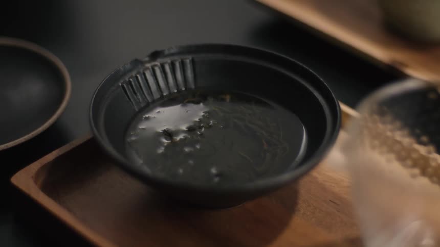 A closeup shot of hot water being poured into a black ceramic bowl containing dark tea leaves which are then covered with a lid and placed on a wooden tray for steeping as part of a traditional ritual