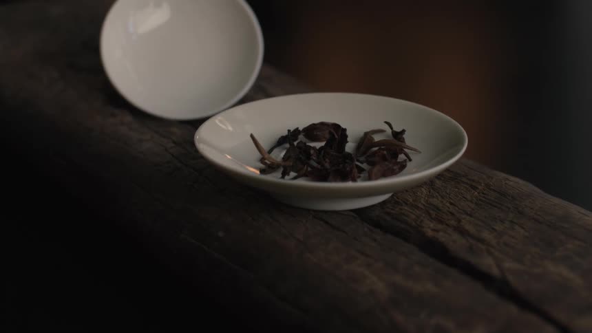 Dried dark tea leaves rest in a shallow white ceramic bowl atop a rough textured wooden beam highlighted by soft contemplative lighting