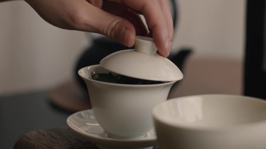The video shows a closeup of a white porcelain teacup which appears to be a gaiwan filled with dry tea leaves on a small saucer a hand gently places the lid onto the cup completing the setting for brewing tea