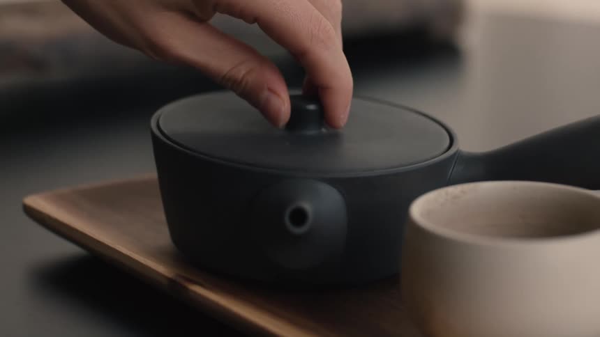 A persons hand reaches in and deliberately lifts the lid from a small dark cylindrical teapot which is sitting on a wooden tray beside a ceramic cup