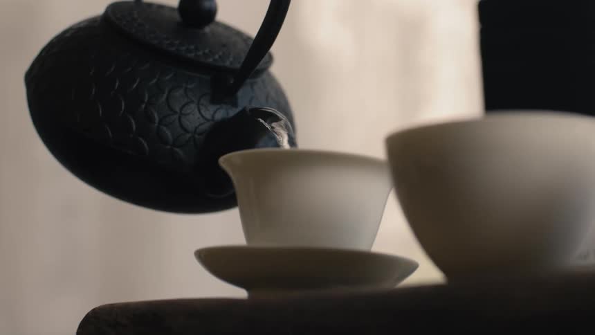 A traditional dark textured castiron teapot is tilted to pour a stream of hot liquid into a small delicate white tea cup the scene focuses on the pouring action which is part of a calming tea preparation ritual