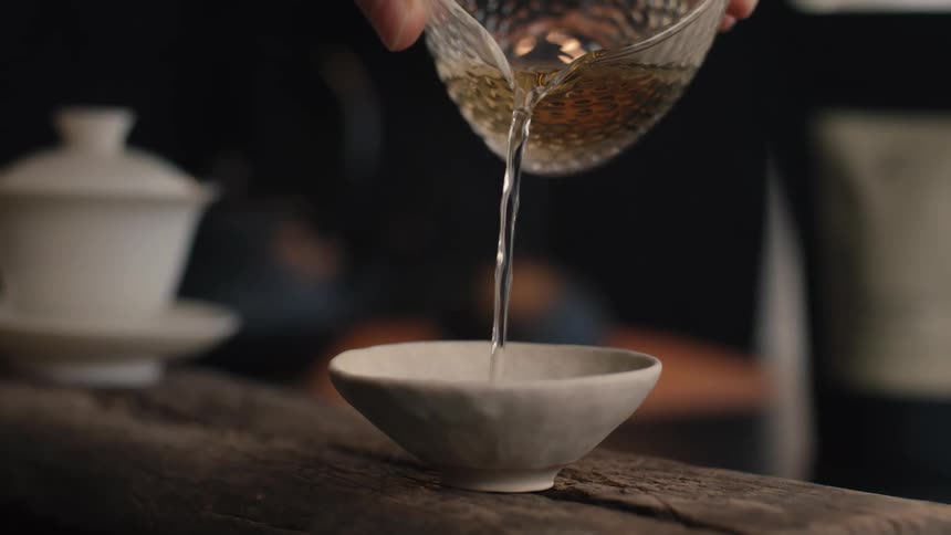 A person pours a stream of light amber liquid from a textured glass pitcher into a small lightcolored ceramic bowl sitting on a rough wooden surface