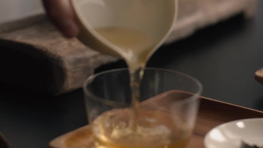 Person pouring tea from a white ceramic jar into a glass cup over a table indoors