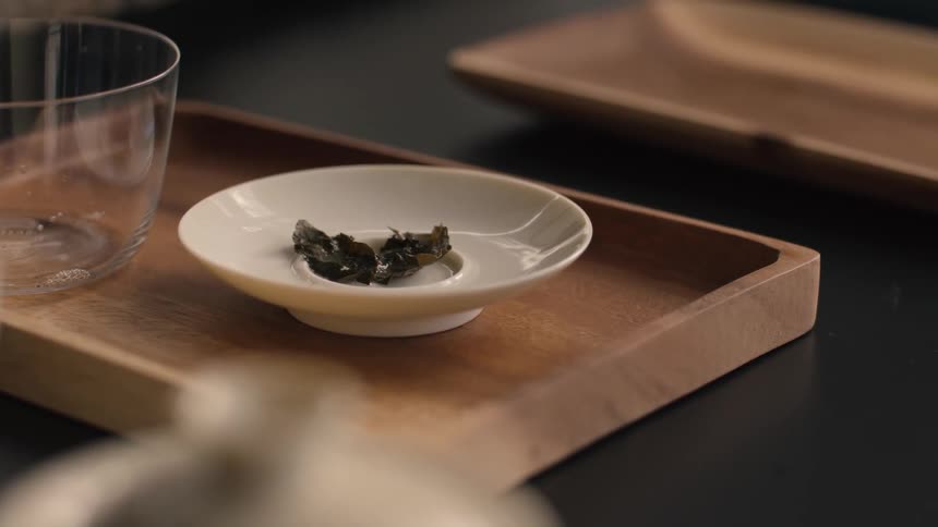 Steeped tea leaves rest in white dish beside glass cup on wooden tray