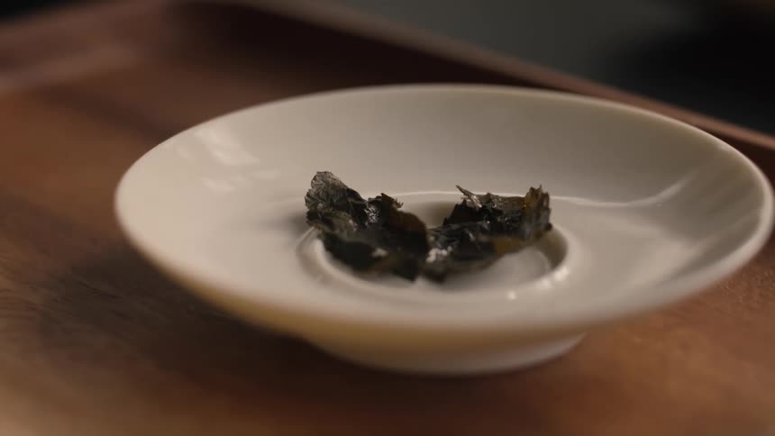 A close view shows two dark green wet tea leaves resting on a small white ceramic dish on a wooden tray