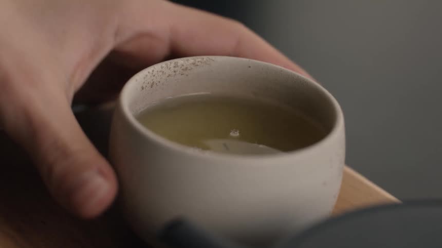 A close view of a hand lifting a small beige ceramic cup filled with a light yellow liquid likely tea from a wooden surface